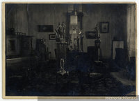 Plano general de un salón: un piano, cuadros, pequeñas esculturas sobre peana, finos muebles, en la ventana, la figura de un hombre junto a una cámara fotográfica.