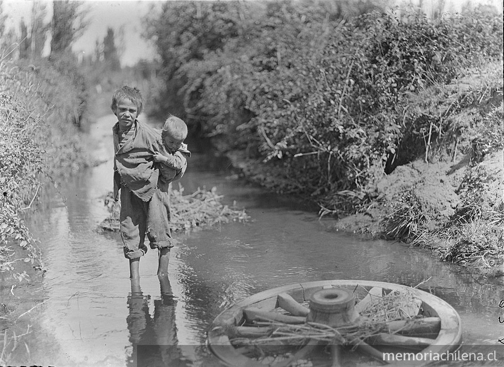 Niño atraviesa un rio con su hemano chico al apa y mira una rueda que flota en las aguas