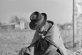 Hombre anciano de manos cruzadas y cabizbajo, con sombrero, sentado sobre un cerro de tierra