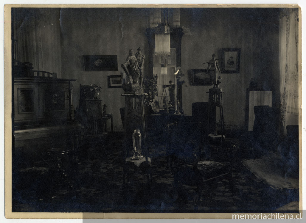 Plano general de un salón: un piano, cuadros, pequeñas esculturas sobre peana, finos muebles, en la ventana, la figura de un hombre junto a una cámara fotográfica.