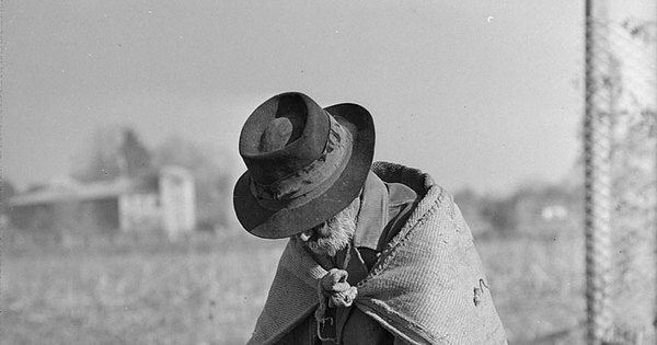 Hombre anciano de manos cruzadas y cabizbajo, con sombrero, sentado sobre un cerro de tierra