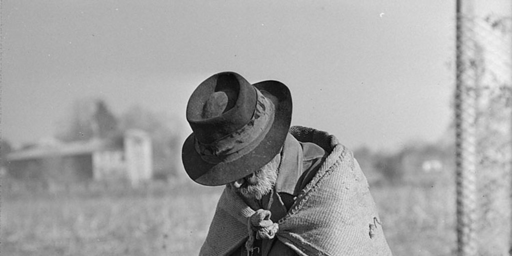 Hombre anciano de manos cruzadas y cabizbajo, con sombrero, sentado sobre un cerro de tierra