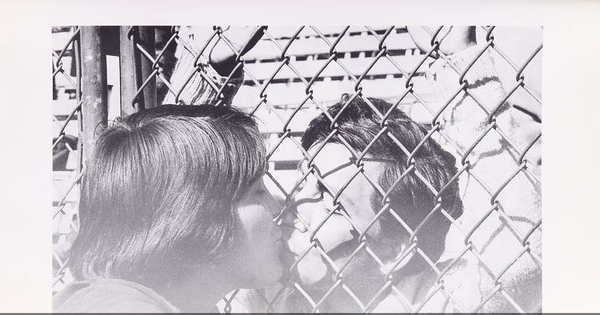 Pie de foto: Día de visita de prisioneros en el Estadio Nacional, 1973. En Cozzi, Adolfo. Estadio Nacional. Santiago: Edit. Sudamericana, 2000. 108 p. Página sin número. Octava foto desde pág. 100, sin pie de foto. Imagen de beso a través de la reja.