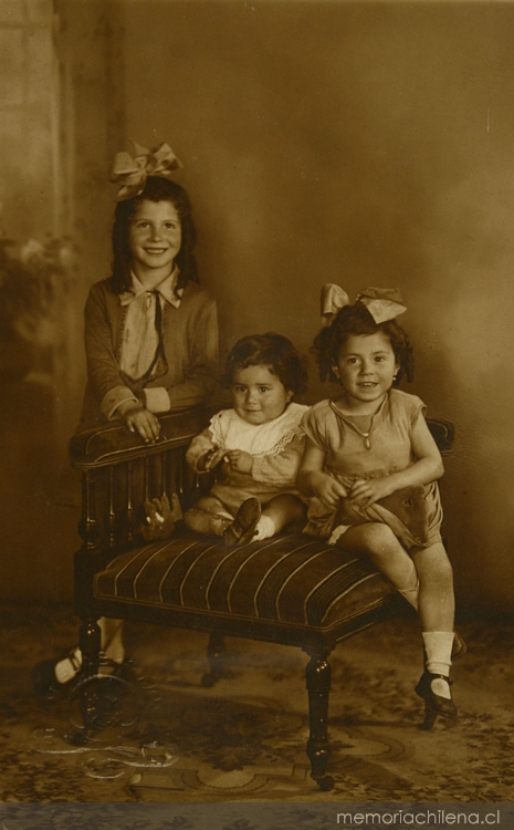 Una niña de pie y las otras dos niñitas sentadas en un sillón, 1925