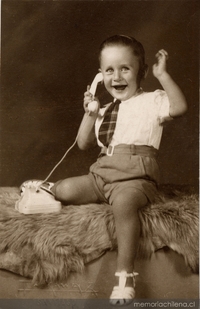 Niño sonriente sentado sobre un felpudo con pantalones cortos, camisa blanca manga corta, corbatín a rayas y chalas blancas, simulando hablar por un teléfono de juguete, 1950