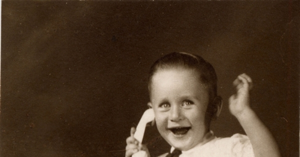 Niño sonriente sentado sobre un felpudo con pantalones cortos, camisa blanca manga corta, corbatín a rayas y chalas blancas, simulando hablar por un teléfono de juguete, 1950