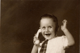 Niño sonriente sentado sobre un felpudo con pantalones cortos, camisa blanca manga corta, corbatín a rayas y chalas blancas, simulando hablar por un teléfono de juguete, 1950