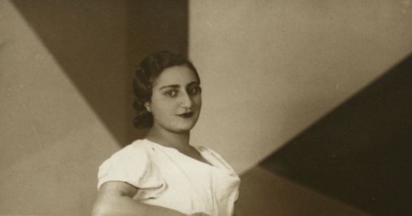 Mujer joven con rasgos árabes, está de pie, con vestido largo de manga corta color blanco, entre 1940 y 1950