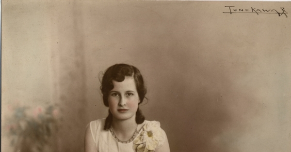 Adolescente de vestido blanco largo adornado con broderí en la parte inferior sentada en un piso de madera sobre alfombra, entre 1930 y 1940