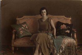 Mujer joven sentada en un sofá, con vestido café y mantilla sobre el regazo, entre 1930 y 1940