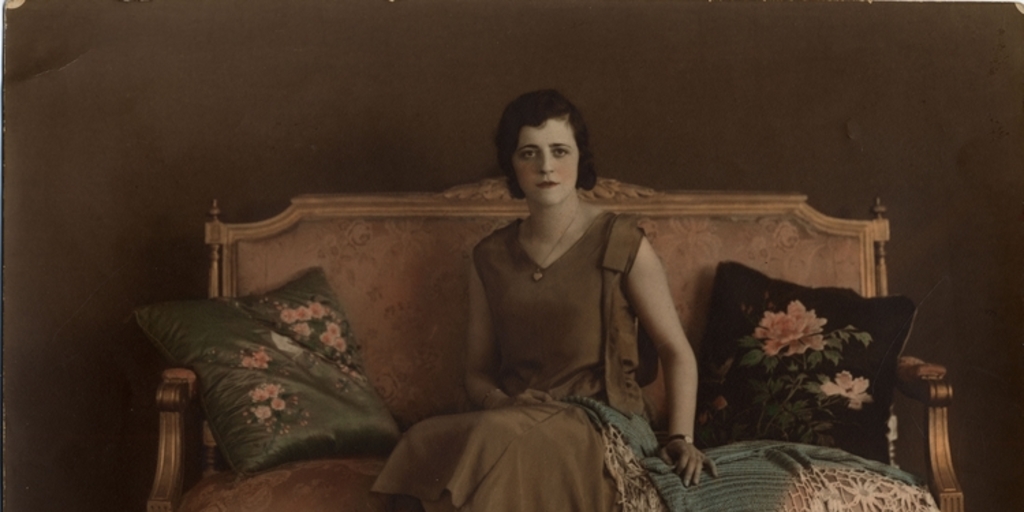 Mujer joven sentada en un sofá, con vestido café y mantilla sobre el regazo, entre 1930 y 1940