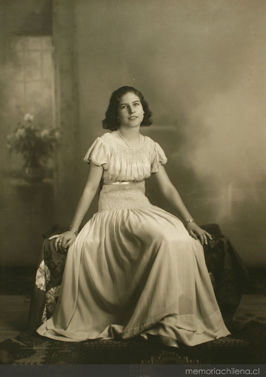 Mujer joven sentada sobre un baúl con un vestido hasta el suelo amplio y acinturado, entre 1928 y 1930