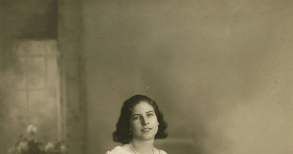 Mujer joven sentada sobre un baúl con un vestido hasta el suelo amplio y acinturado, entre 1928 y 1930