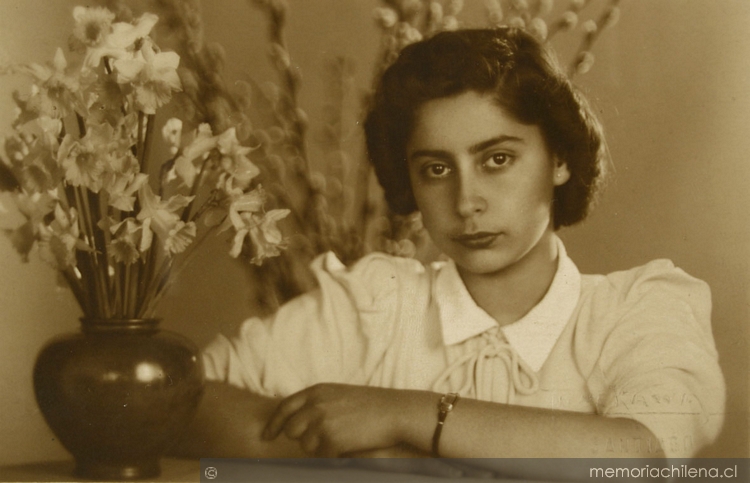 Mujer joven, vestida de verano y apoyada en un mueble: en frente tiene un macetero con flores blancas y a sus espaldas se ve unos brotes de ramas largas, entre 1940 y 1950